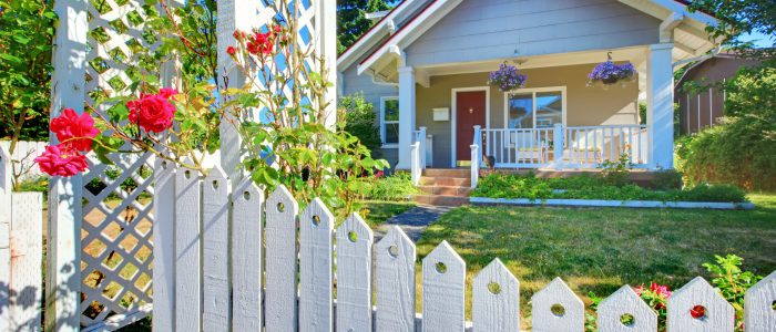 House tucked behind a beautiful white picket fence and green grass and flowers.